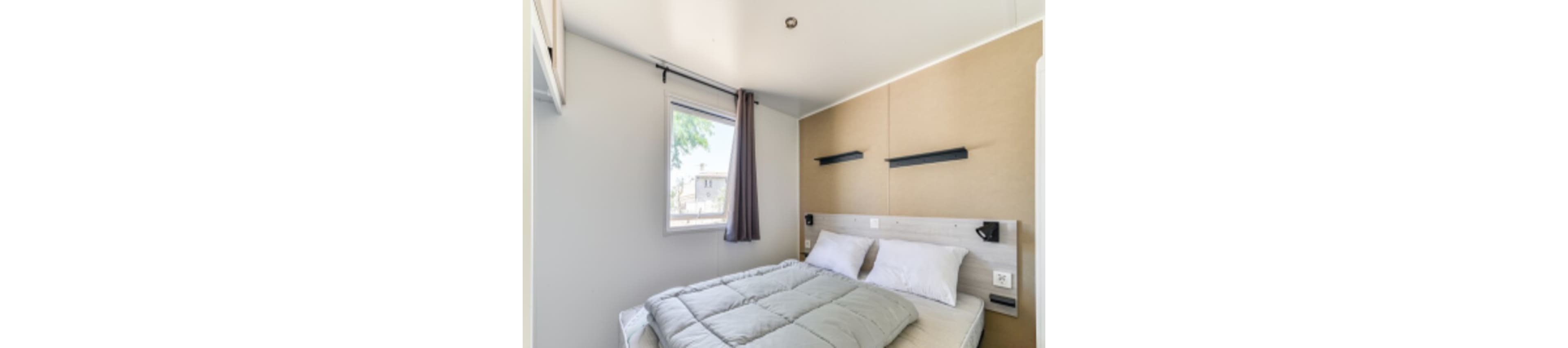 A compact, modern bedroom features a neatly made double bed with white and grey bedding. The room has a single window with a grey curtain, allowing natural light to brighten the space. The walls are painted in neutral tones, and two floating shelves are mounted above the bed. The overall style is minimalist and clean, with a calm and inviting atmosphere.