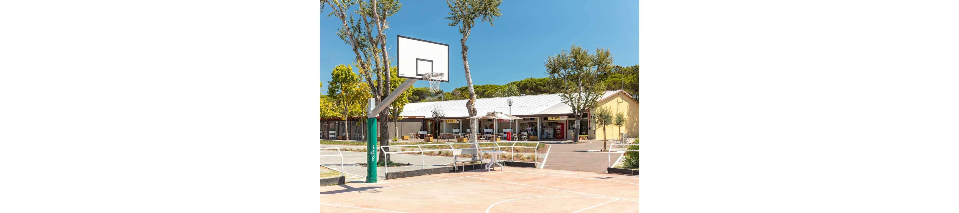 An outdoor basketball court stands in the foreground with a single hoop and backboard. Behind the court, a small row of cafes or shops with outdoor seating is visible under a bright, clear sky. Tall trees provide some shade and greenery around the paved area. The scene suggests a relaxed recreational space in a warm, sunny climate.