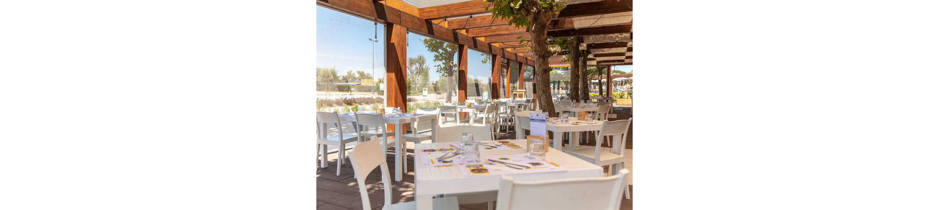 An open-air restaurant terrace features neatly arranged white tables and chairs under a wooden pergola. Sunlight filters through the beams and surrounding trees, creating a warm and inviting atmosphere. Tables are set with placemats, glasses, and menus, ready for guests to dine. Large windows and open sides reveal greenery and a bright blue sky in the background.