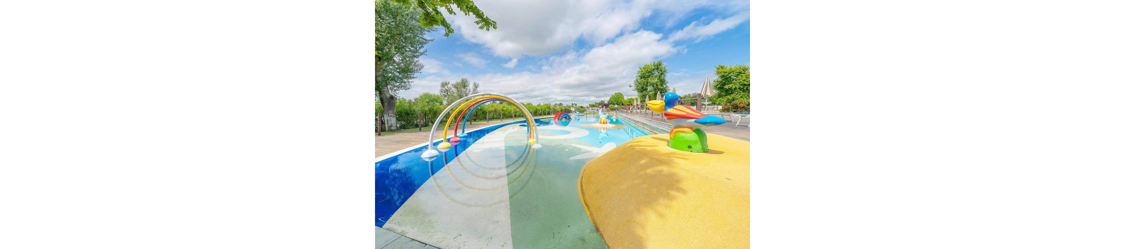 An outdoor splash pad features shallow water and colorful play structures. Rainbow-shaped spray arches and animal-themed fountains are arranged along the pool. Bright yellow and blue surfaces contrast with the clear sky and green trees in the background. The scene suggests a family-friendly recreational area on a sunny day.