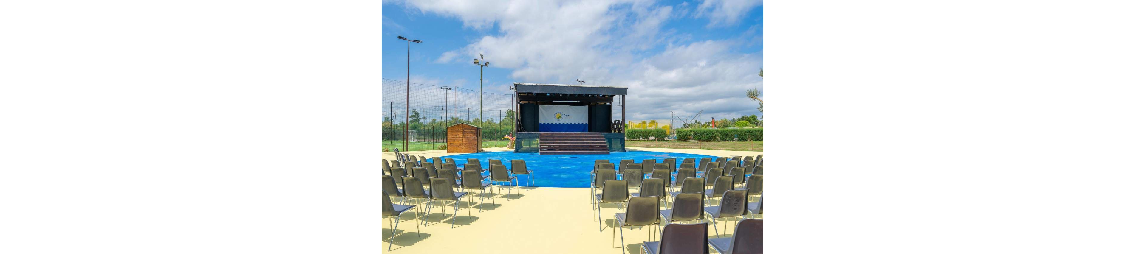 An outdoor performance stage is set up in a sunny open area with rows of empty chairs facing it. The stage features a simple backdrop with a blue and white wave design and a circular sun graphic. Surrounding the venue are green hedges, sports nets, and tall light poles under a partly cloudy sky. The scene suggests a venue prepared for a show, presentation, or community event.