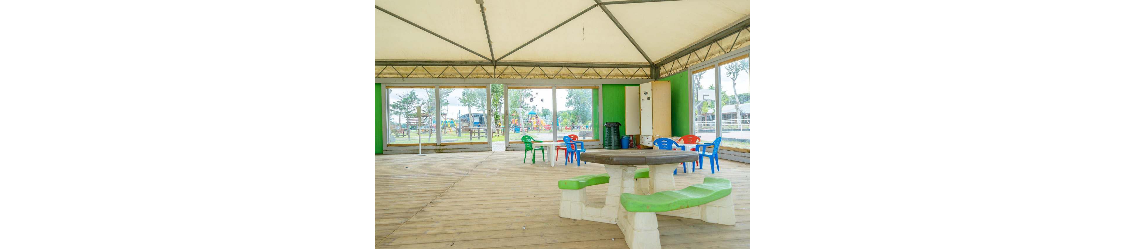 A bright indoor pavilion features a round plastic table with green seats in the foreground. Colorful plastic chairs are scattered around the wooden floor, suggesting a space for children. Large glass windows reveal an outdoor playground with slides and other play structures. The overall scene feels open, airy, and family-friendly.
