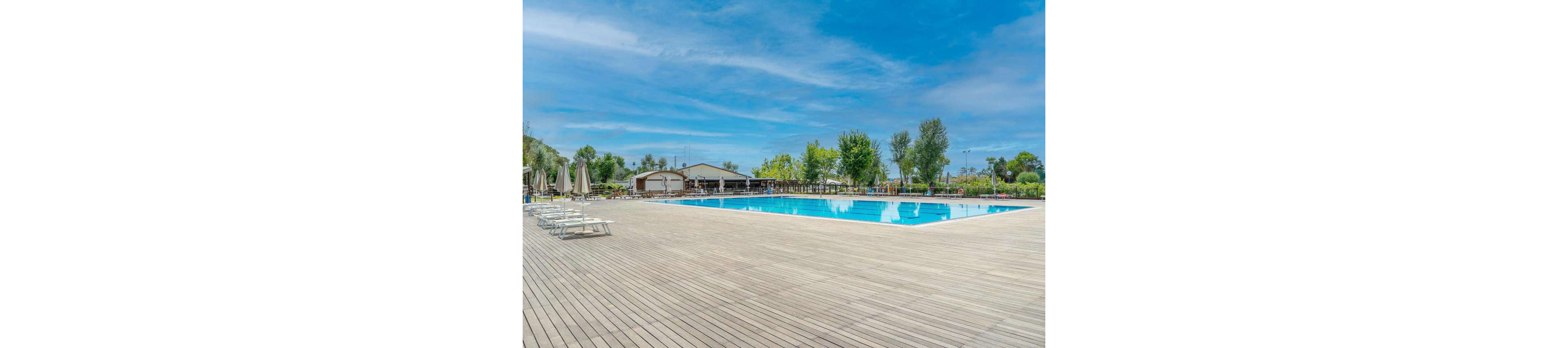 A large rectangular outdoor swimming pool sits in the middle of an expansive wooden deck. Closed umbrellas and empty lounge chairs line one side of the pool, suggesting a quiet moment between guests. In the background, a low building and lush green trees frame the leisure area under a bright blue sky with light clouds. The scene conveys a calm, sunny day at a resort or recreational facility.