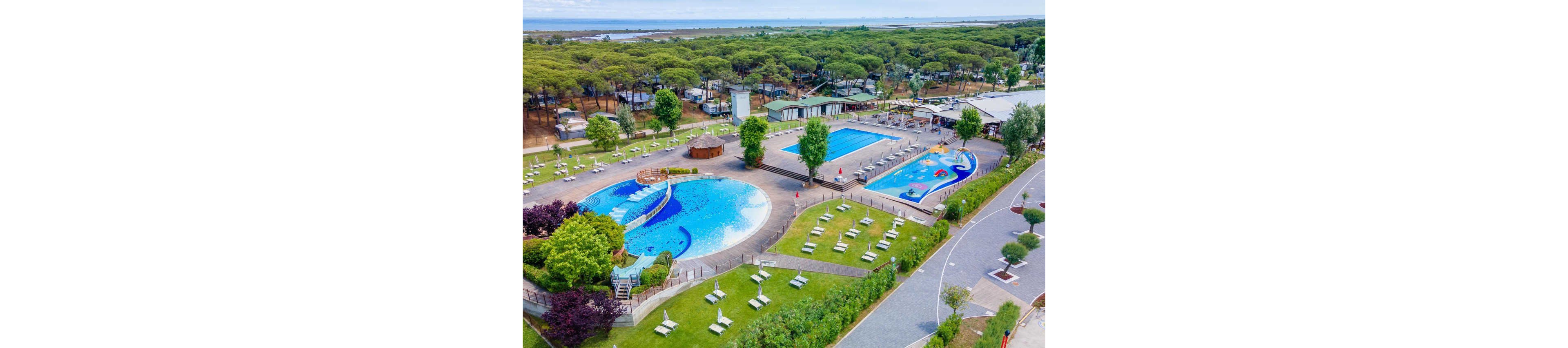 A large outdoor resort complex features multiple swimming pools surrounded by green lawns and trees. The main circular pool has a playful, colorful design with slides and shallow areas for children. Nearby, a long rectangular lap pool and another themed pool with bright graphics provide additional swimming options. Sun loungers are neatly arranged on the grass, and pathways connect the pool area to surrounding buildings and a forested campground in the background.