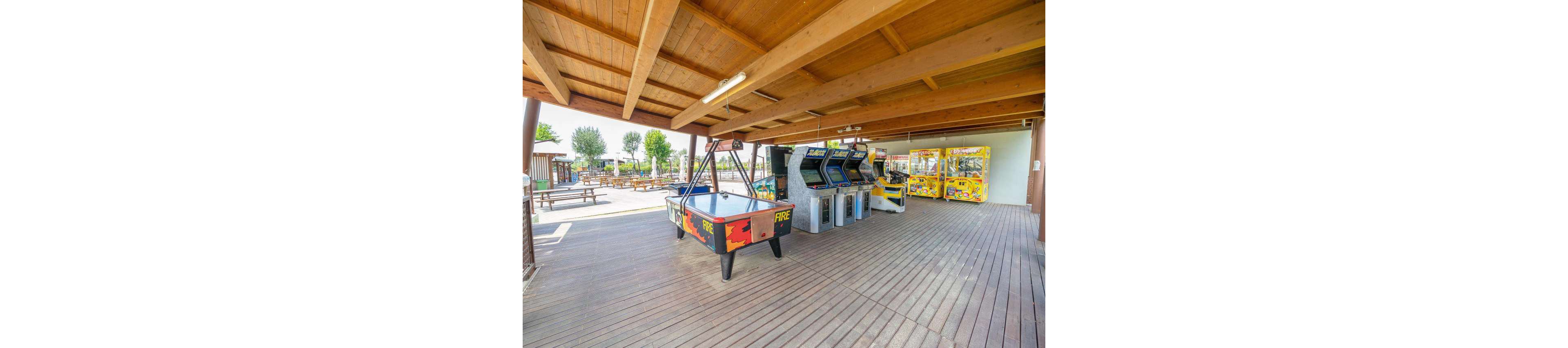 An open-air arcade area features several classic arcade game machines and two air hockey tables under a wooden roof. The space has a wide wooden deck floor and is partially open to the outside. In the background, picnic tables and trees suggest a leisure or recreation park setting. Bright colors on the machines and tables create a playful, family-friendly atmosphere.
