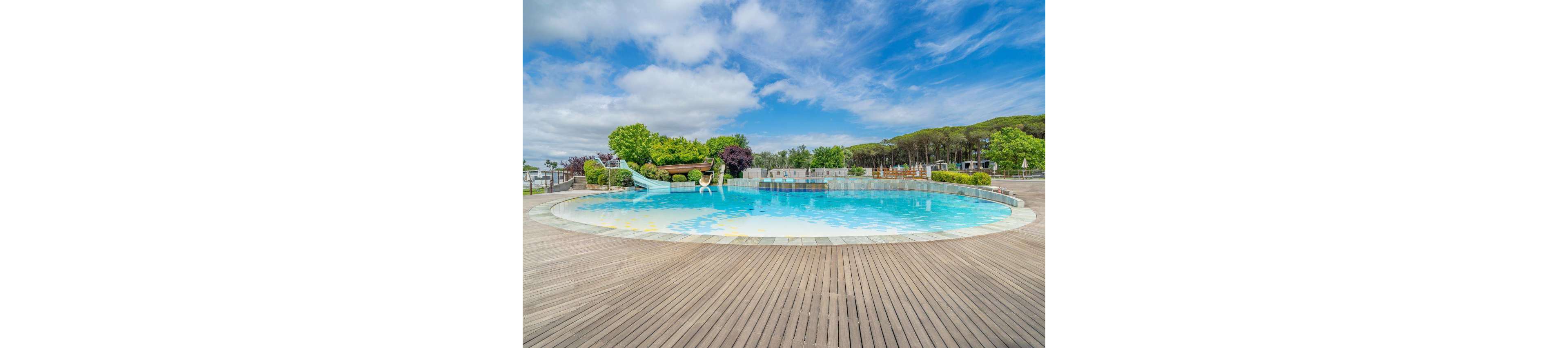 A large circular outdoor swimming pool sits in the center of a spacious wooden deck. The pool features a curved water slide and a lifebuoy hanging on the railing. Lush green trees and landscaped shrubs surround the area, with a forested hillside in the background. A bright blue sky with scattered clouds adds to the calm, vacation-like atmosphere.