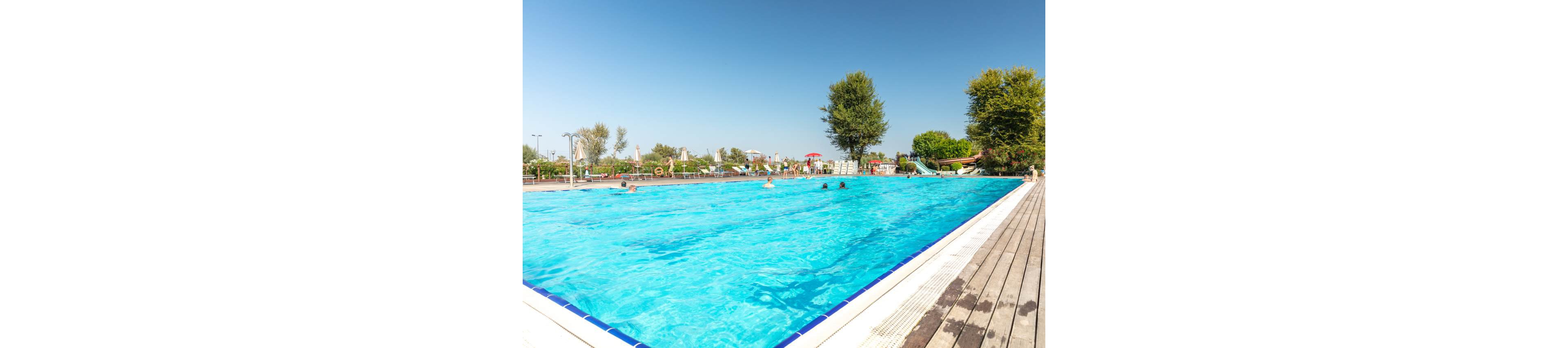 A large outdoor swimming pool sparkles under a clear blue sky. Several people are swimming and relaxing in the bright turquoise water. Trees, umbrellas, and a waterslide are visible in the background, suggesting a family-friendly recreation area. A wooden deck runs along the poolside, adding a warm natural contrast to the cool water.