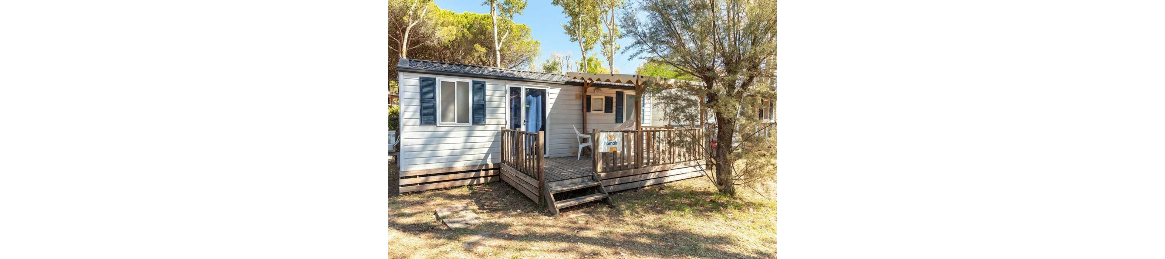 A small mobile home cabin with a wooden deck is set among tall trees in a sunny outdoor area. The structure features light-colored siding, blue shutters, and a simple front porch with railings. A single white plastic chair sits on the deck near the entrance. The surrounding environment includes dry grass, scattered shade, and a relaxed, rustic campsite atmosphere.