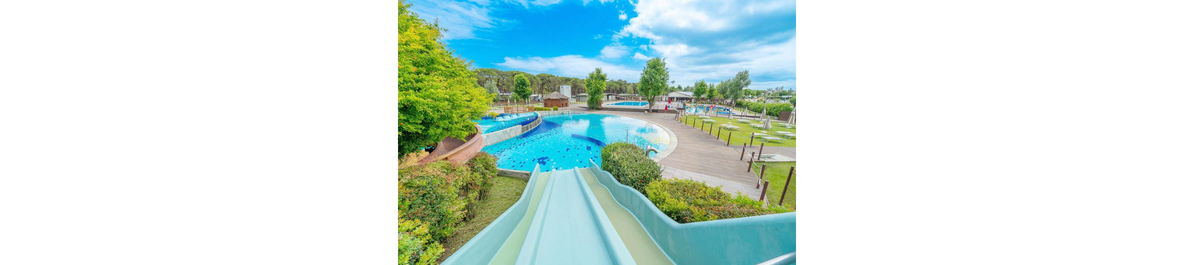 An outdoor water park features a large curving slide leading into a bright blue swimming pool. Multiple pools and winding water channels are surrounded by green bushes, trees, and wooden walkways. Sun loungers and umbrellas are arranged on the grass and deck areas, suggesting a family-friendly leisure setting. The sky is partly cloudy, adding a fresh and inviting atmosphere to the scene.