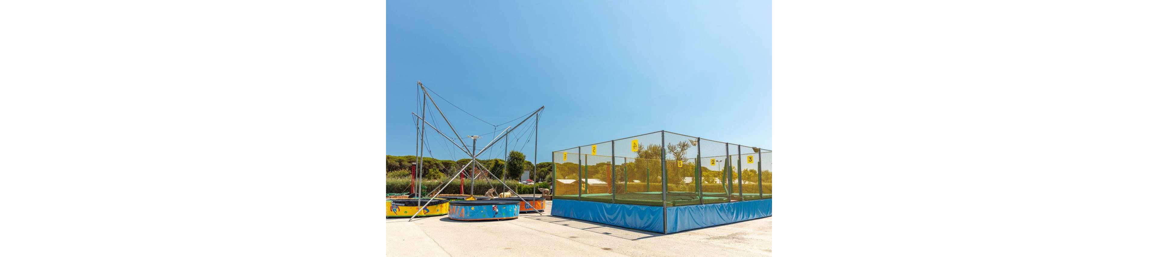 An empty outdoor amusement area features a bungee trampoline structure beside a large enclosed trampoline court. The scene is set on a sunny day with clear blue skies and distant green trees. Bright yellow safety nets and blue padding add vibrant color to the recreational setup. No people are present, emphasizing the equipment and open space.