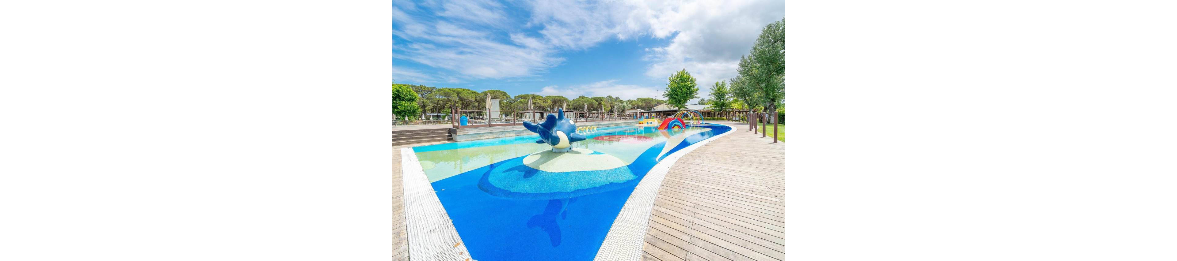 An outdoor children’s splash pool features a playful dolphin slide in shallow water. The pool is surrounded by wooden decking and shaded seating areas in the background. Bright colors, including blue, yellow, and a rainbow arch, create a fun and inviting atmosphere. Trees and a partly cloudy sky frame the family-friendly water play area.