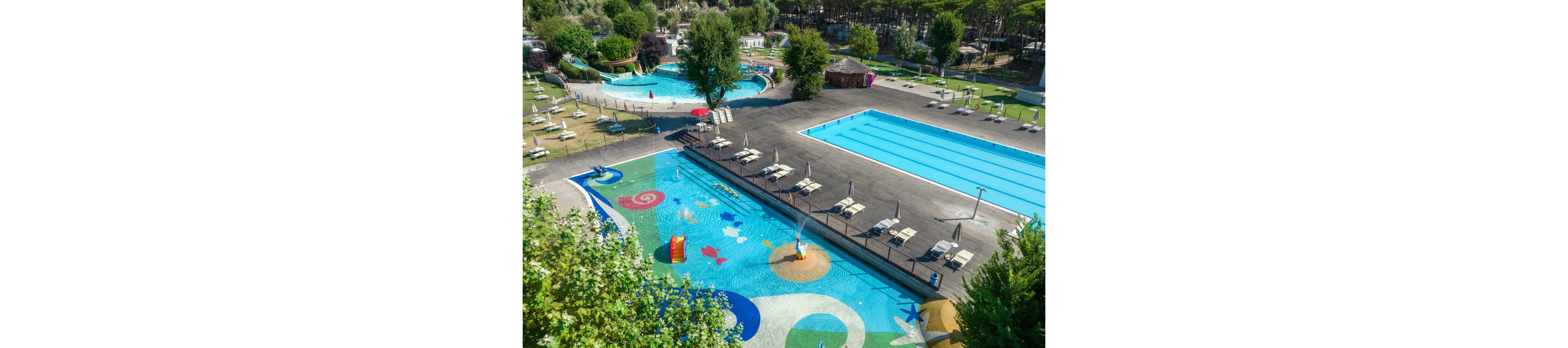 A vibrant outdoor water park features several swimming pools of different sizes, including a colorful shallow kids’ pool with playful graphics and small slides. Sun loungers and umbrellas are neatly arranged around the pools, suggesting a relaxing recreational area. The surrounding landscape includes trees, grassy areas, and pathways that create a resort-like atmosphere. The scene is bright and sunny, emphasizing leisure, summer fun, and family-friendly activities.