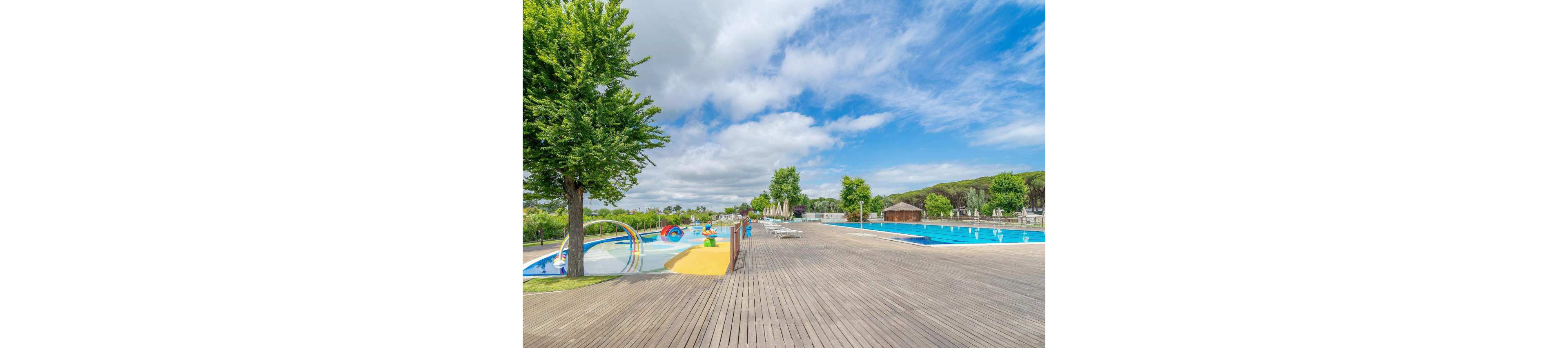 A large outdoor swimming pool stretches alongside a wide wooden deck under a partly cloudy sky. On the left, a shallow children's splash area features colorful water play structures and slides. The scene is surrounded by green trees and low hills, giving the area a relaxed, natural atmosphere. Lounge chairs and umbrellas are visible in the distance, suggesting a family-friendly vacation resort.