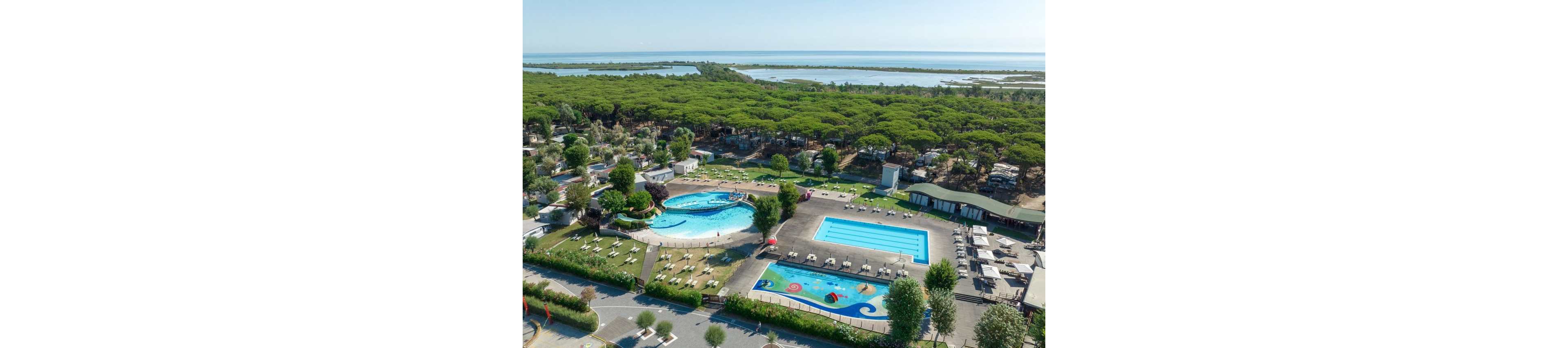 A large holiday resort features multiple outdoor swimming pools surrounded by sun loungers and umbrellas. The scene is captured from above, showing a circular pool, a rectangular lap pool, and a children’s splash area with colorful play elements. The resort is bordered by dense green forest that stretches toward a calm coastal lagoon and the sea in the distance. Buildings, pathways, and parked vehicles indicate a well-developed vacation destination in a natural seaside setting.
