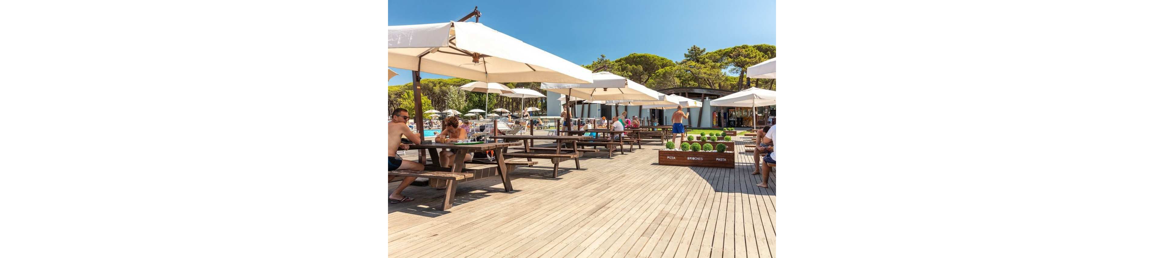 An open-air beach bar features wooden picnic tables under large white umbrellas on a spacious deck. People relax, eat, and socialize in swimwear, enjoying the sunny weather near the coast. In the background, a modern low building houses the bar area, surrounded by lush green trees. Decorative planters with green spheres and text add visual interest along the edge of the terrace.