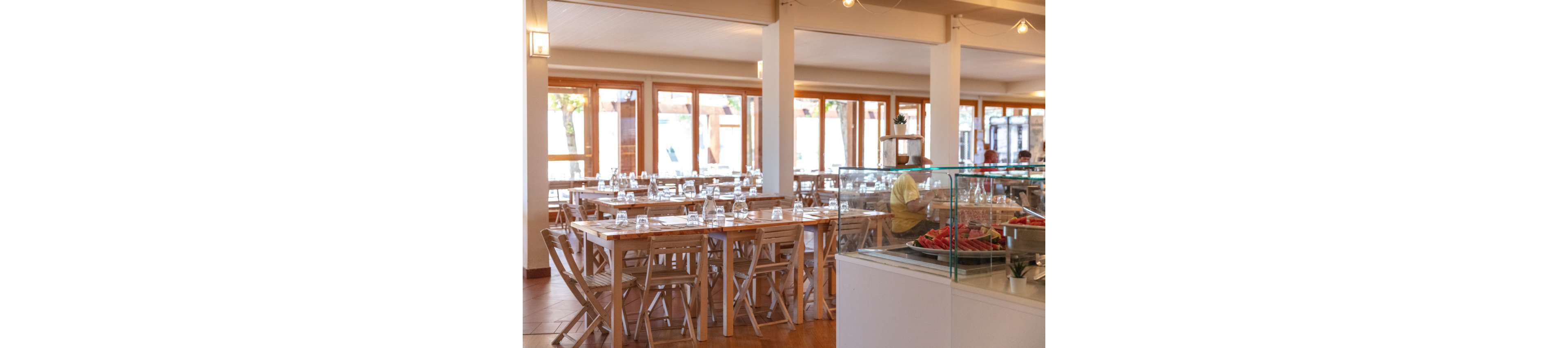 A spacious restaurant dining area features rows of wooden tables and chairs neatly set with upside-down drinking glasses. Large windows along the walls let in abundant natural light, creating an airy and welcoming atmosphere. In the foreground, a glass display counter shows plates of sliced watermelon and other food items. A single seated person in a yellow shirt is visible near the back, adding a sense of quiet, off-peak service time.