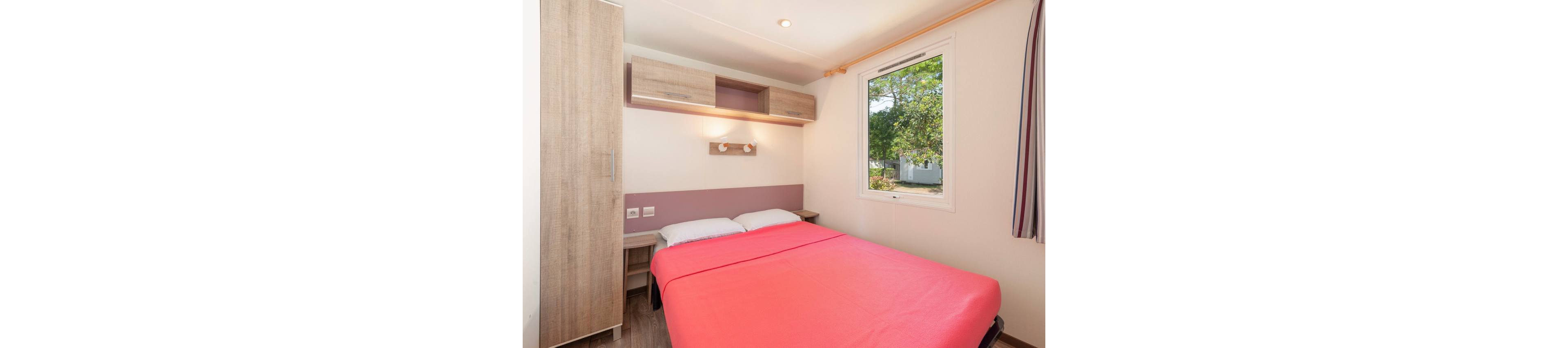 A compact bedroom features a double bed covered with a bright pink bedspread and white pillows. Light wood furniture, including a tall wardrobe and overhead cabinets, adds storage along the wall. A large window lets in natural light and offers a view of green trees outside. The room has a clean, minimalist design with soft pastel accents and striped curtains on one side.