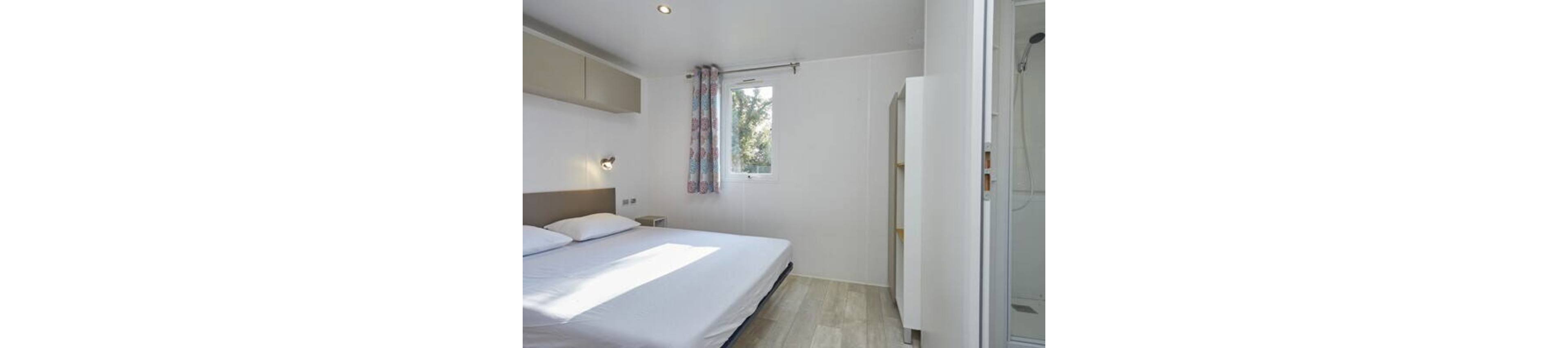 A small modern bedroom features a double bed with white linens against a light-colored wall. Natural light enters through a single window with patterned curtains, casting soft shadows on the floor. Built-in cabinets and shelves provide simple storage along the walls. A glass shower enclosure is visible to the right, suggesting an en-suite layout in a compact living space.