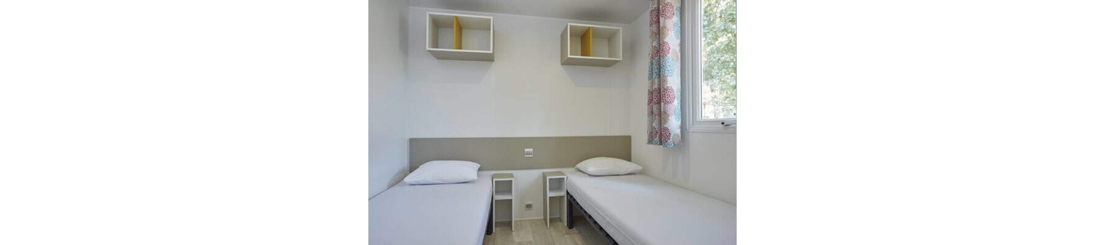 The image shows a small bedroom with two single beds placed parallel along opposite walls. Each bed has a plain white pillow and mattress, emphasizing a clean and minimalist look. Between the beds are two narrow white bedside shelves, and above them are wall-mounted cube shelves with yellow dividers. A window with colorful patterned curtains lets in natural light, brightening the otherwise neutral room.