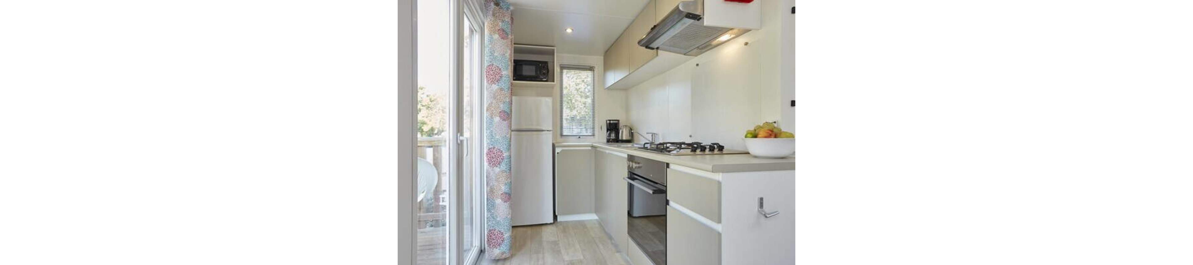 A narrow modern kitchen features light cabinetry and a compact layout. A refrigerator and microwave stand at the far end beside a small window. The countertop holds a kettle, coffee maker, and a bowl of fresh fruit near a gas cooktop and built-in oven. Sliding glass doors with patterned curtains open to an outdoor balcony, allowing natural light to fill the space.