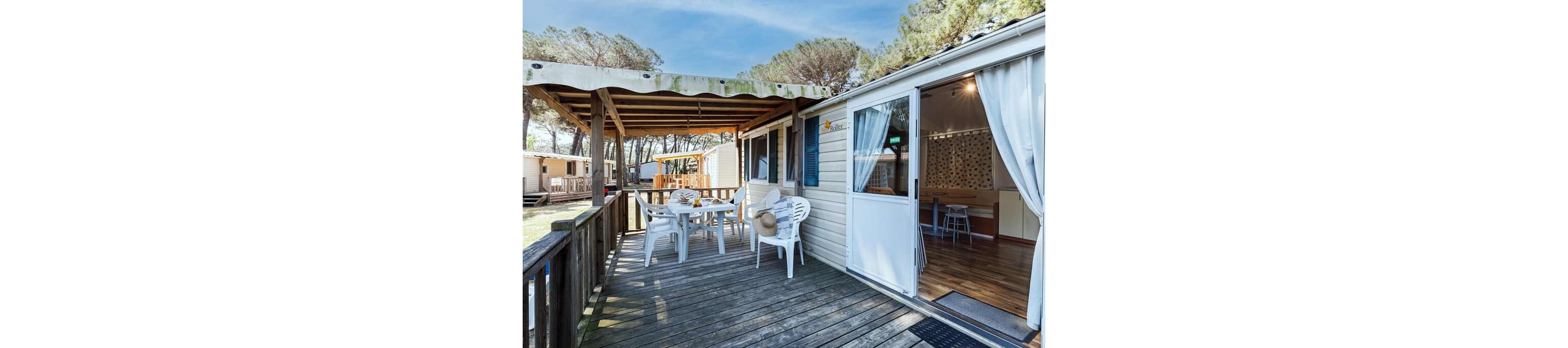 A wooden deck terrace extends from a light-colored mobile home set in a forested campsite. White plastic chairs and a table are arranged under a partial awning, with a sun hat resting on one of the chairs. The sliding door is open, revealing a bright interior with wooden flooring and a small dining area. Other similar cabins and tall trees are visible in the background, suggesting a relaxed holiday setting.