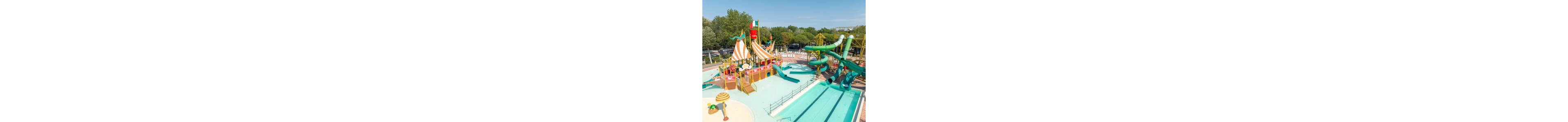A vibrant pirate-themed water play structure stands beside a large outdoor pool. Multiple green and orange slides twist down from towers and a ship-like platform with striped sails. The surrounding area includes lounge chairs, shallow splash zones, and playful decorative elements under a clear blue sky. Trees and parked cars frame the background, emphasizing the park’s open-air setting.
