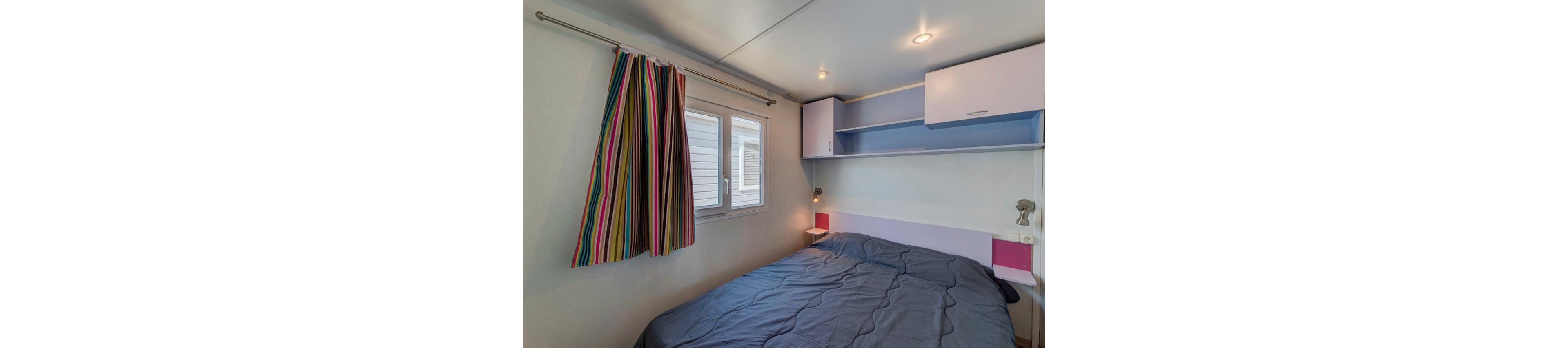 Compact bedroom interior featuring a double bed with a dark blue comforter. The room includes a window with bright multicolored striped curtains that add a playful touch. Built-in shelves and cabinets above the bed provide storage and are finished in white and pastel colors. Recessed ceiling lights and small wall-mounted reading lamps create a warm, inviting atmosphere.