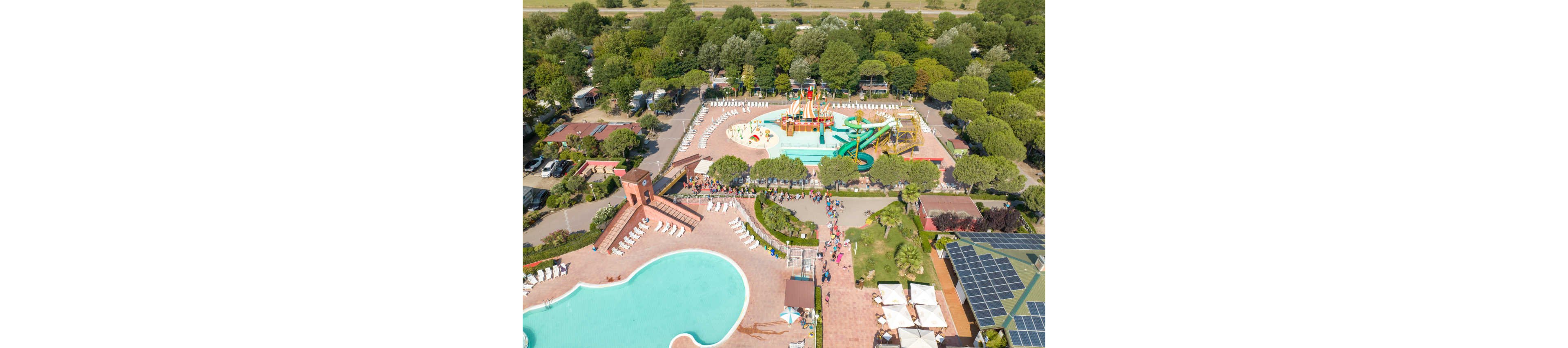A large outdoor water park resort is captured from above, showing multiple pools and colorful water slides surrounded by trees. One pool has a playful splash area with slides and water features, while another larger pool curves around a sunbathing deck lined with loungers. Guests are visible walking along pathways and gathering near the attractions, creating a lively family atmosphere. The surrounding area includes landscaped greenery, buildings, and solar panels, emphasizing a spacious holiday setting.
