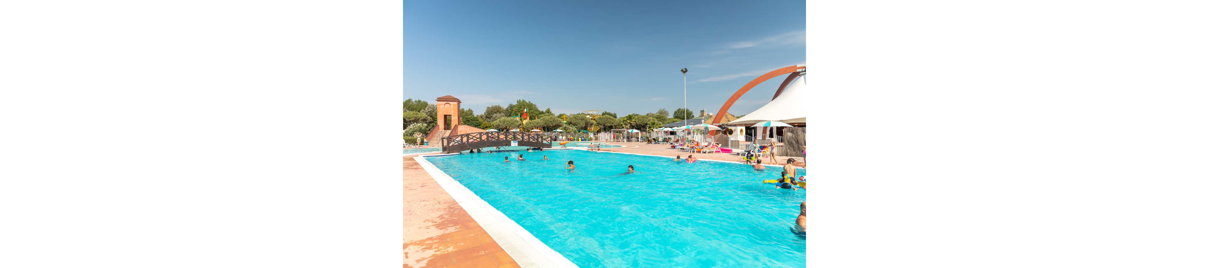 An outdoor swimming pool is filled with people enjoying a sunny day at a resort. Children and adults play in the bright blue water while others relax on loungers around the pool. A small wooden bridge and a tower-like structure add visual interest to the poolside area. In the background, umbrellas, trees, and clear blue sky create a cheerful vacation atmosphere.