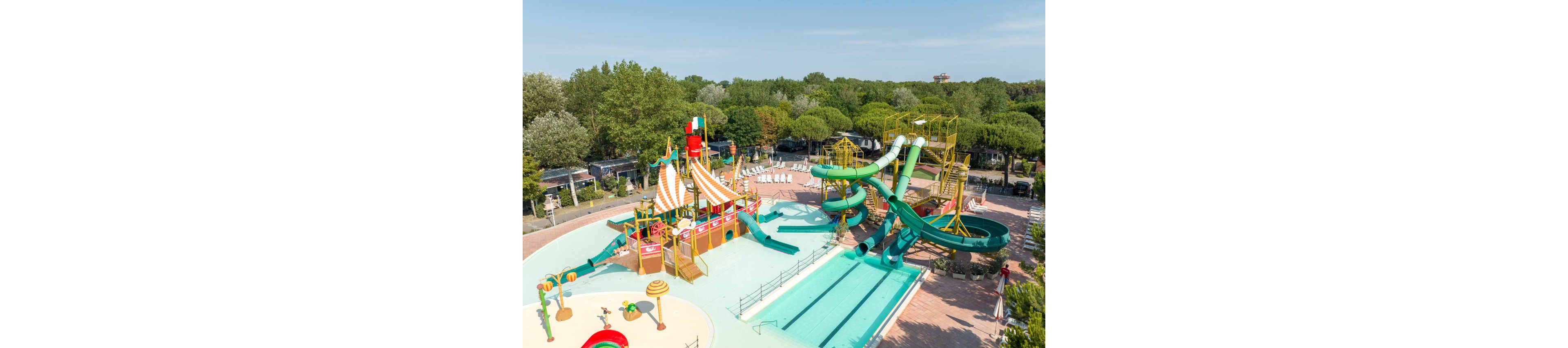 An outdoor water park features a large pirate-ship themed play structure with multiple slides and water features. Adjacent to it are tall twisting green water slides that empty into a long rectangular pool. The area is surrounded by lounge chairs, paved walkways, and lush green trees under a clear blue sky. The scene suggests a family-friendly recreational space designed for summer fun and relaxation.