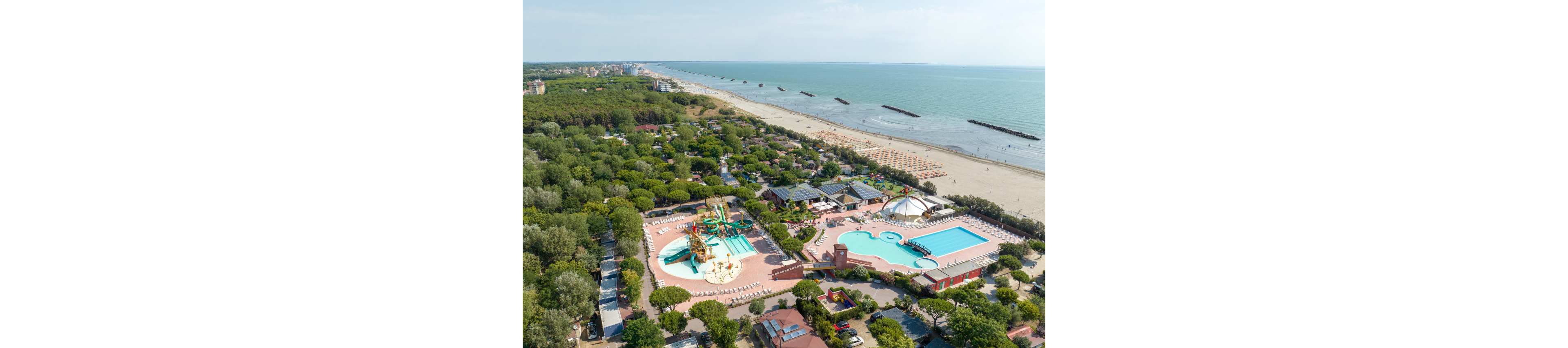 A large seaside resort complex is captured from above, showing multiple swimming pools and water slides near a sandy beach. The resort is surrounded by dense green trees and numerous low-rise buildings and campsites. Sun loungers and umbrellas line the shoreline, while breakwaters extend into the calm sea. The overall scene suggests a popular holiday destination with family-friendly water attractions.