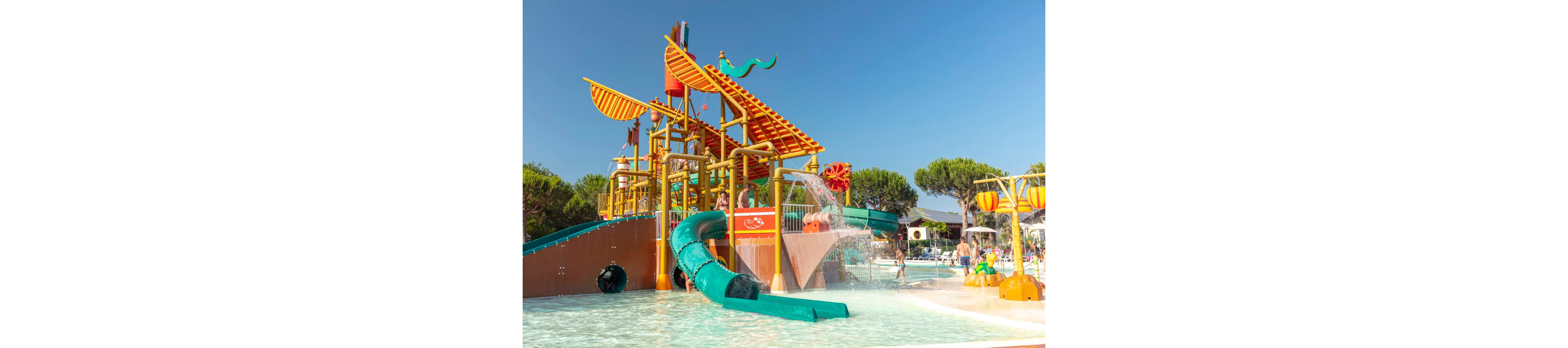 A vibrant outdoor water playground features a large structure with multiple slides and splash elements. Bright orange and yellow platforms rise above a shallow pool, with a prominent green tube slide curving into the water. Children and families enjoy the sunny day, playing among fountains and water jets. Tall trees and clear blue sky frame the fun, resort-like setting.