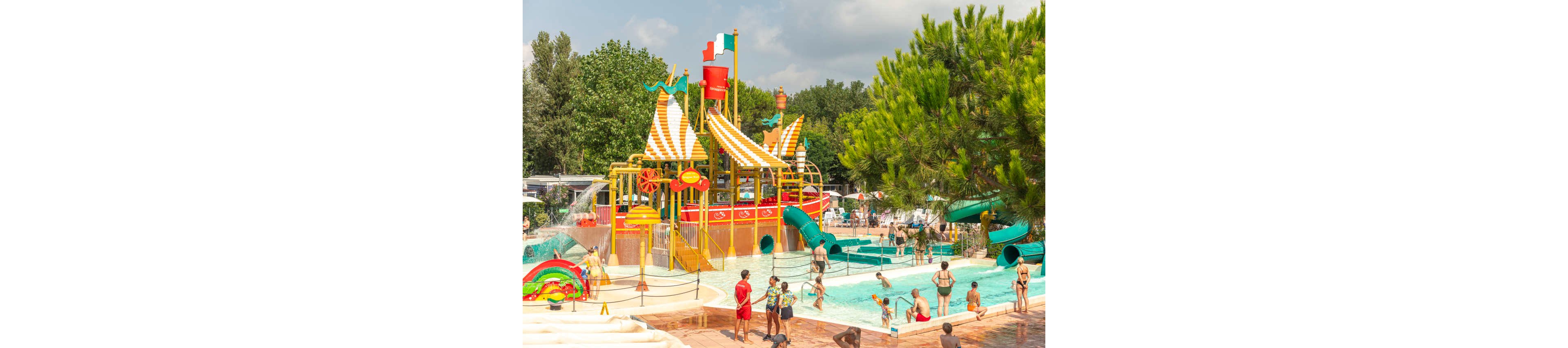 A vibrant pirate-themed water play structure stands in the middle of a busy outdoor pool. Children and adults enjoy slides, fountains, and shallow water under a sunny sky. Bright red, yellow, and green elements create a playful, colorful atmosphere. Trees and nearby facilities surround the aquatic area, giving the scene a lively holiday resort feel.