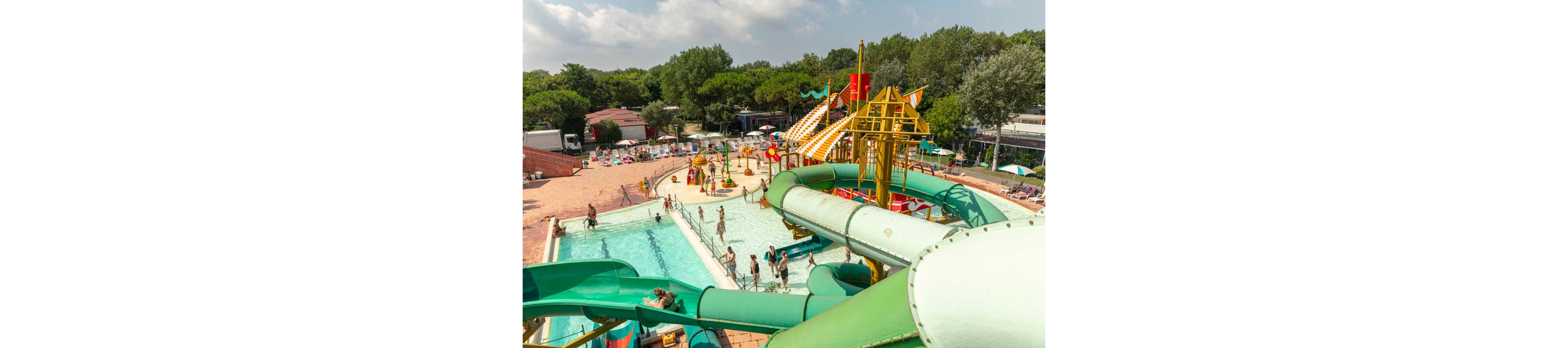 An outdoor water park features large green and yellow water slides emptying into bright blue pools. Families and children play in the shallow water and walk along the poolside deck under the summer sun. In the background, more visitors relax on lounge chairs and under umbrellas near trees and park buildings. The overall scene feels lively, fun, and family-friendly.