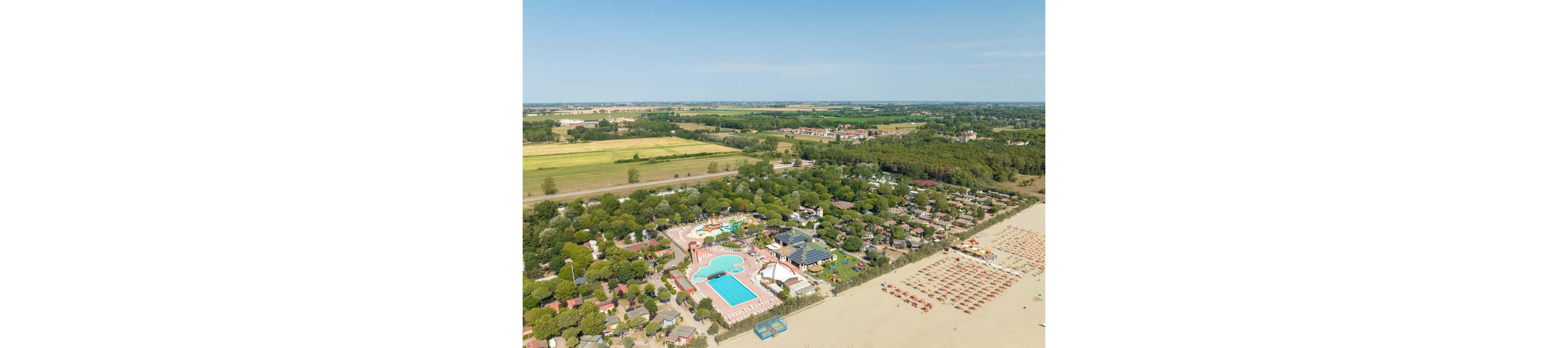 A large seaside holiday resort is captured from above, showing pools, buildings, and tree-lined plots. The complex sits directly beside a wide sandy beach filled with rows of organized sun loungers and umbrellas. Beyond the resort, green fields and scattered houses stretch into the distance under a clear blue sky. The image highlights the contrast between the structured vacation area and the surrounding rural landscape.