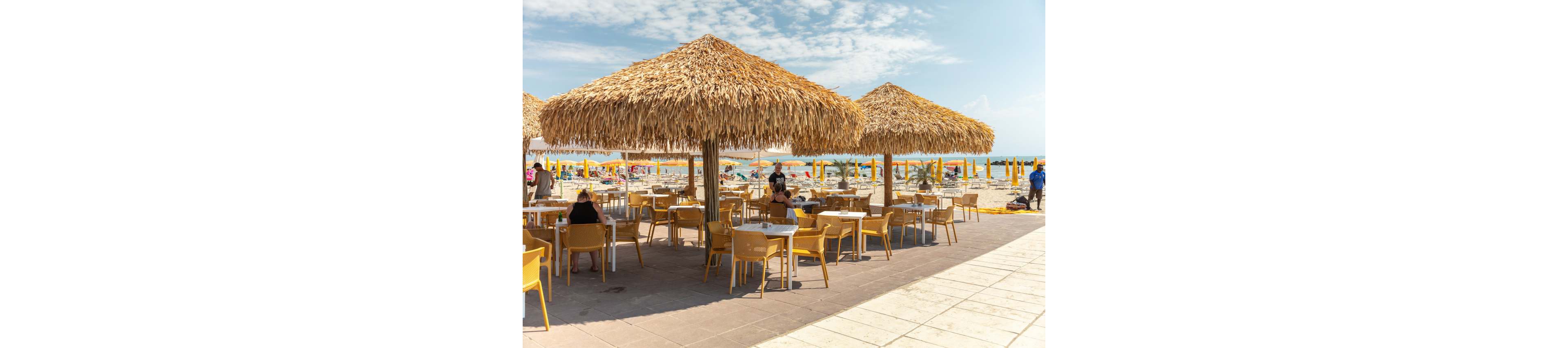 Outdoor café tables sit beneath large straw parasols on a sunny beachfront promenade. Yellow chairs and umbrellas create a bright, summery atmosphere near the sea. People relax at the tables and on the sandy beach in the background. The sky is partly cloudy, adding texture to the coastal scene.