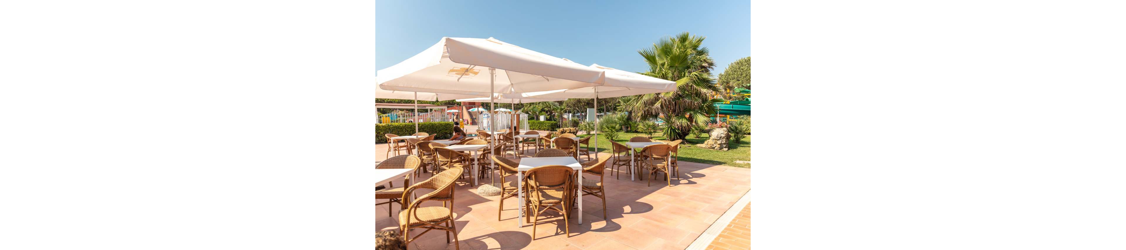 An outdoor cafe terrace features several wicker chairs and white tables arranged under large white sun umbrellas. The seating area is set on a tiled patio adjacent to a green garden with palm trees and manicured shrubs. In the background, colorful playground structures and fencing suggest a family-friendly environment. The bright sunlight and clear blue sky create a warm, summery atmosphere.
