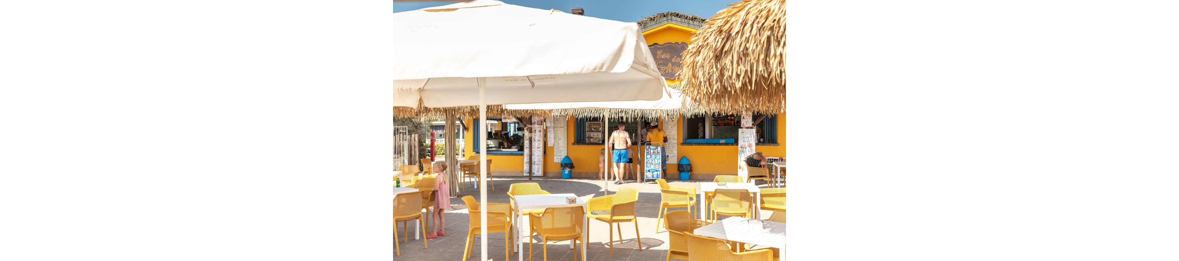 An outdoor beach bar with bright yellow walls and furniture sits under large white umbrellas. Several people in swimwear stand near the counter while others relax at empty tables on the terrace. Straw-thatched roofs and sunlit tiles emphasize the coastal, vacation setting. The scene conveys a warm, relaxed summer atmosphere by the sea.