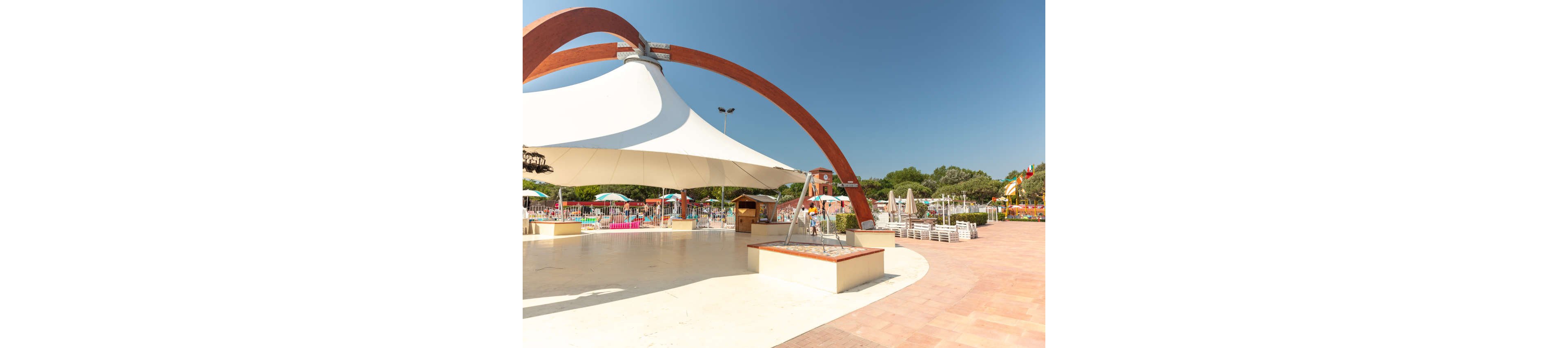 An expansive outdoor leisure area features a large white tensile shade canopy supported by a curved red arch. In the background, colorful umbrellas and sun loungers surround a swimming pool complex. The scene is bright and sunny with clear blue skies and green trees framing the space. Flags and small structures suggest a holiday resort or water park environment.