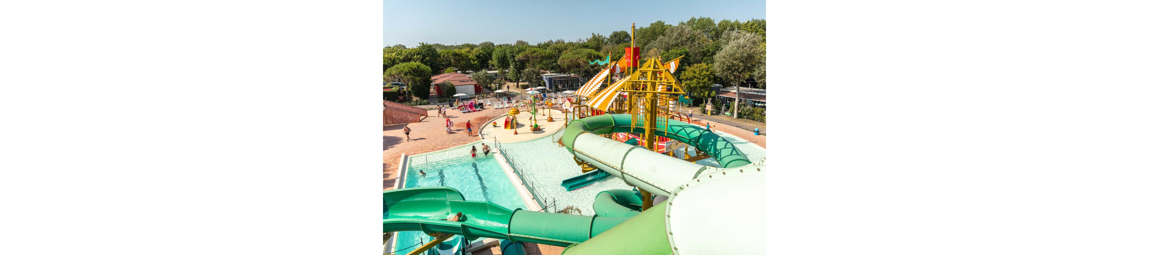 An outdoor water park features multiple large green and yellow slides leading into bright blue pools. Families and children enjoy swimming and playing in the shallow water under sunny skies. The scene includes lounge chairs, umbrellas, and surrounding trees that create a relaxed vacation atmosphere. The overall setting suggests a busy summer day at a resort or campground aquatic area.