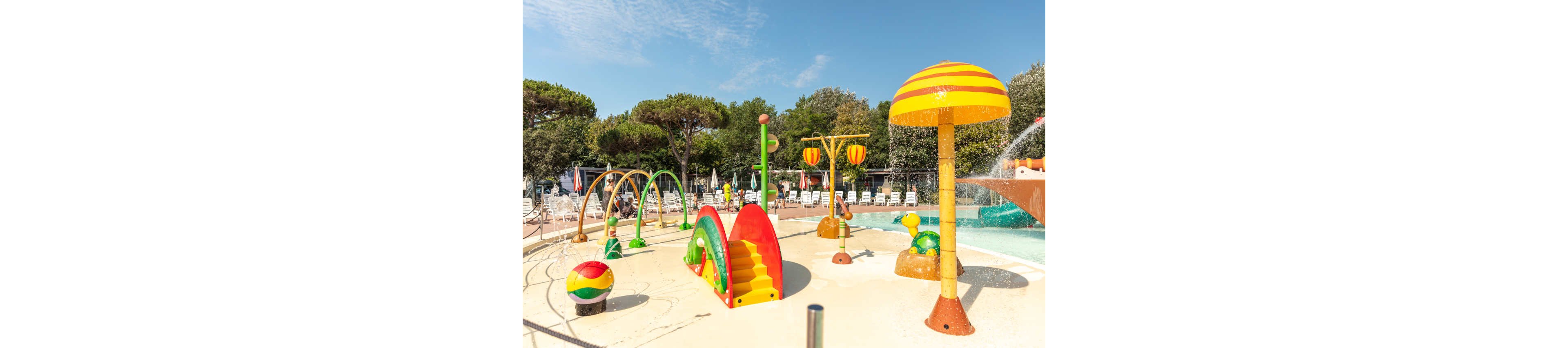 Brightly colored water play structures stand in a shallow splash area at an outdoor pool. The scene includes slides, arches, and mushroom-shaped fountains spraying water. Lounge chairs and umbrellas line the background, with trees providing a natural backdrop. The sunny weather and vivid colors create a cheerful, family-friendly atmosphere.