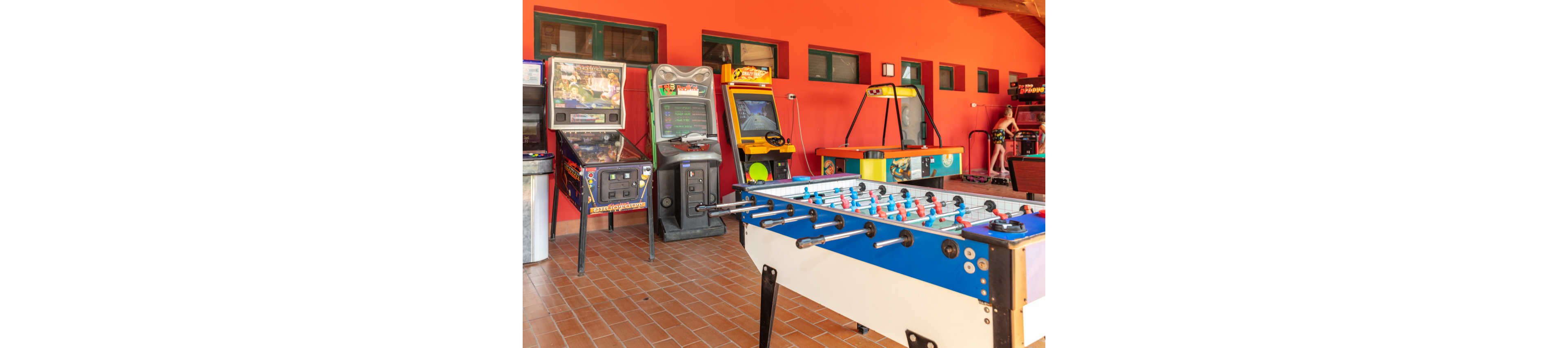 A bright indoor arcade room features a foosball table in the foreground. Several classic arcade and pinball machines line the red-painted wall behind it. The floor is covered with terracotta-colored tiles, and natural light enters through rectangular windows. A person is seen in the background near one of the arcade machines, adding a casual, playful atmosphere.