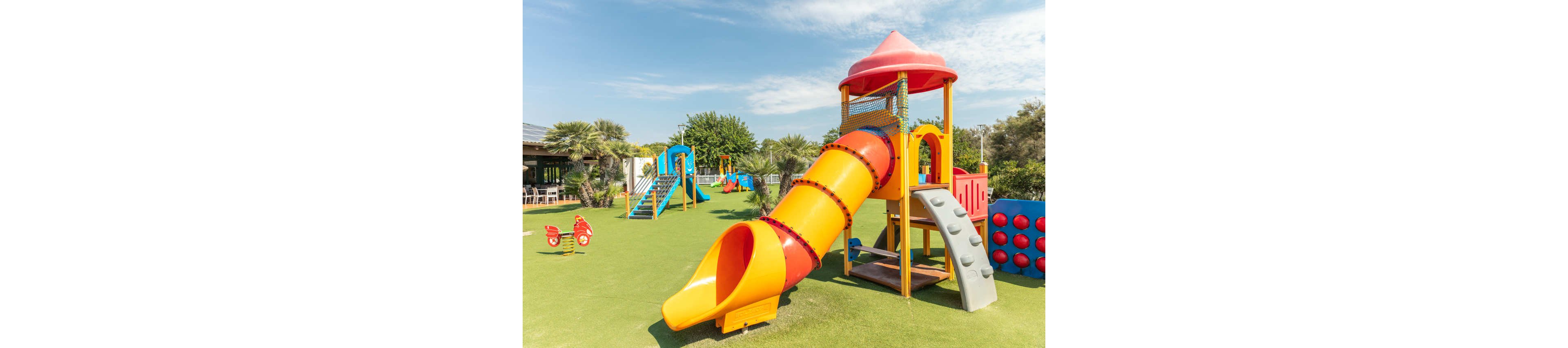 A bright outdoor playground features a large yellow and orange tube slide with a red roofed tower. The play area is set on green artificial grass under a sunny blue sky with scattered clouds. Additional equipment such as climbing structures, swings, and spring riders are visible in the background. Palm trees and a nearby building suggest the playground is part of a park or resort setting.