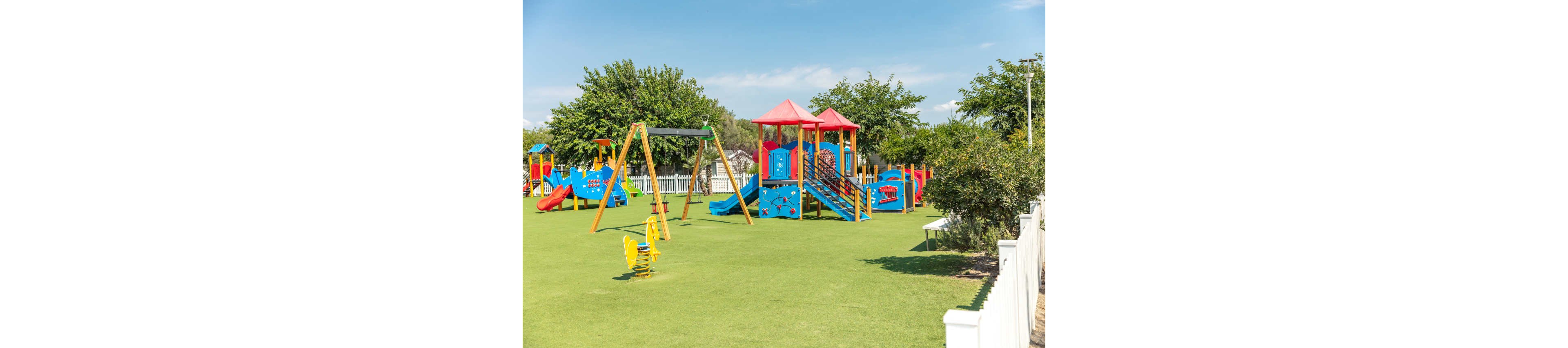 A bright, colorful playground stands on a large grassy field under a clear blue sky. The play area features slides, climbing structures, and swings in red, yellow, and blue. White fences and green trees surround the playground, creating a safe and inviting environment. The scene appears calm and empty, emphasizing the equipment and open space.