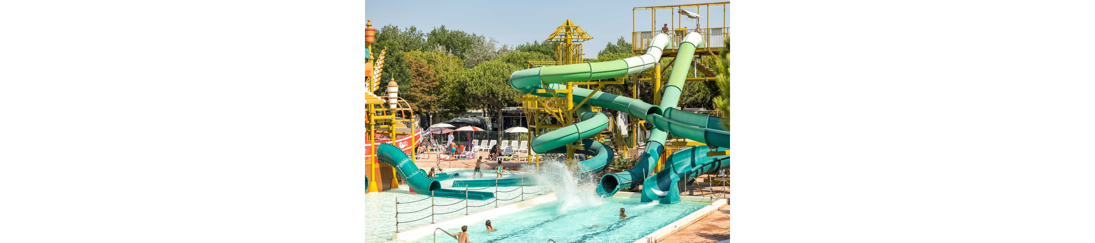 Large twisting green and turquoise water slides empty into a bright outdoor pool. Children and families play in the water and wait near the slide exits under sunny skies. Yellow slide supports and nearby lounge chairs suggest a busy water park setting. Trees and additional play structures frame the background, creating a lively summer atmosphere.