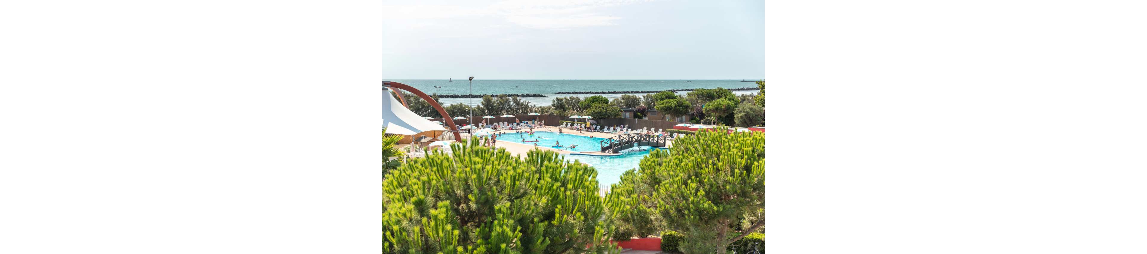 An outdoor swimming pool at a seaside resort is surrounded by sun loungers and umbrellas. People relax and swim in the bright blue water under a clear sky. Lush green trees and shrubs frame the pool area in the foreground. In the background, the calm sea stretches to the horizon with breakwaters visible along the shoreline.