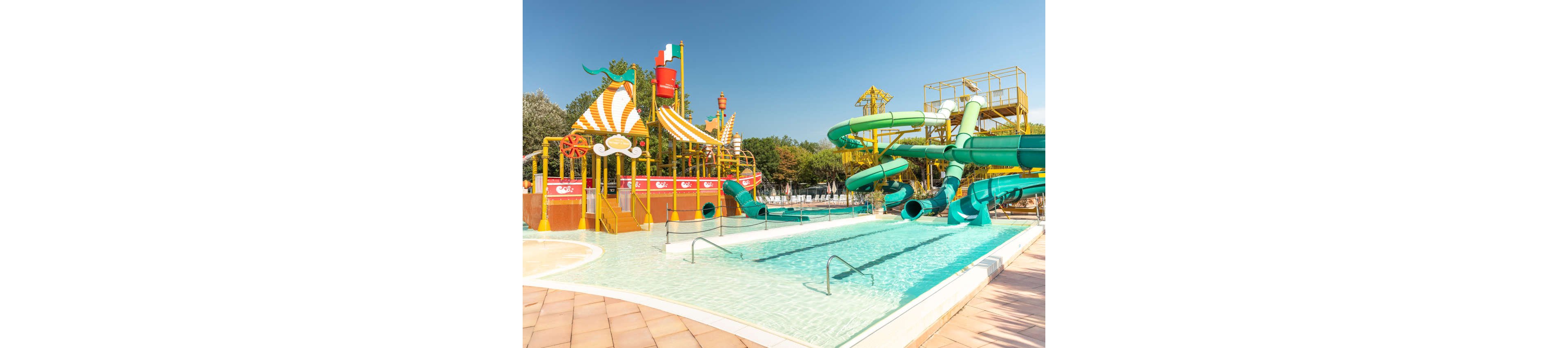 A bright outdoor water park features multiple twisting green water slides emptying into a shallow pool. On the left, a large play structure with striped canopies, stairs, and water features adds to the fun atmosphere. The pool area is calm and empty, with clear blue water and metal handrails leading into the shallow sections. Trees and additional park structures are visible in the background under a clear blue sky.