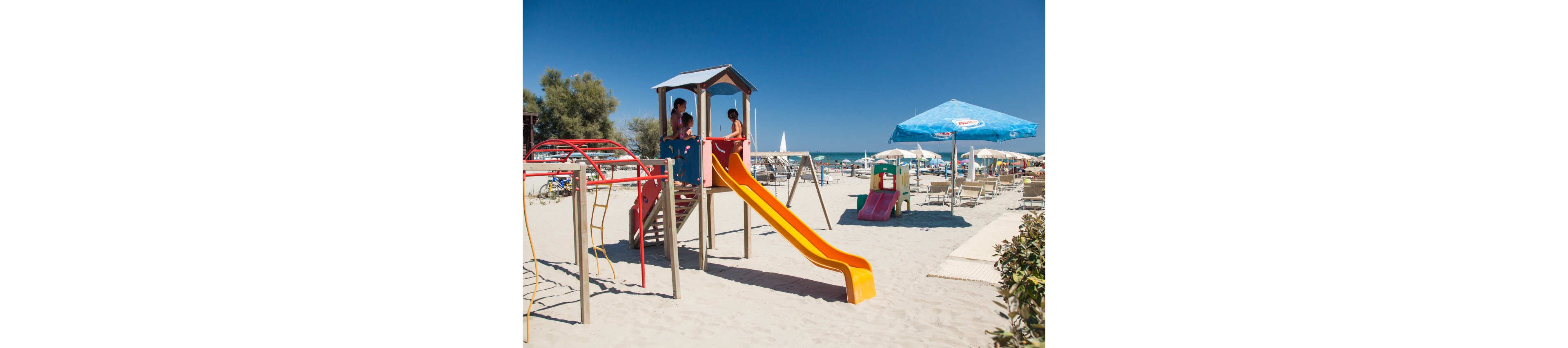 Children play on a bright yellow slide and climbing structure set directly on a sandy beach. Sun loungers and umbrellas are arranged in the background near the shoreline, with the blue sea and clear sky beyond. A large blue parasol and additional small play equipment add more color to the scene. The overall atmosphere is sunny, relaxed, and family friendly.