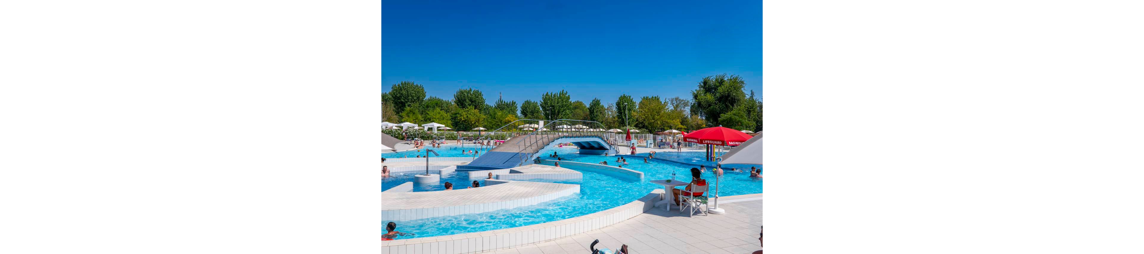 An outdoor swimming pool complex is filled with people enjoying a sunny day. Adults and children swim and wade in the bright blue water while others relax on the poolside deck. A curved pedestrian bridge crosses over the pool, adding visual interest to the scene. Red umbrellas and green trees in the background create a vibrant summer atmosphere under a clear blue sky.