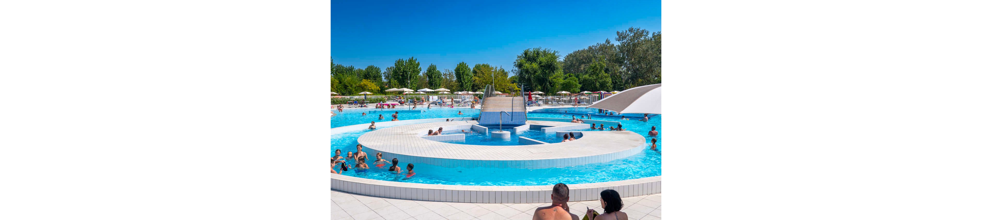 An outdoor swimming pool is filled with people relaxing and playing in the water. A circular central island with steps and a small structure creates a focal point in the pool. Sun loungers and umbrellas line the background under a clear blue sky, with trees providing a natural backdrop. The scene suggests a warm summer day at a public pool or resort, with families and children enjoying leisure time together.